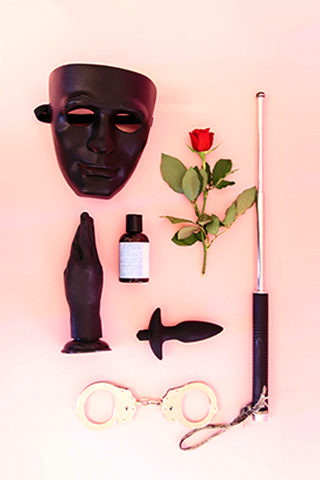 A series of kinky tools, such as a mask, a silicone hand, a pair of metal handcuffs, an anal plug, a riding crop, and a bottle of lubricant sit atop a pink background with a red rose.