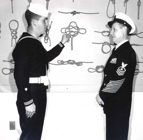 Black and white picture of an enlisted sailor pointing at rope knots on the wall in front of an officer