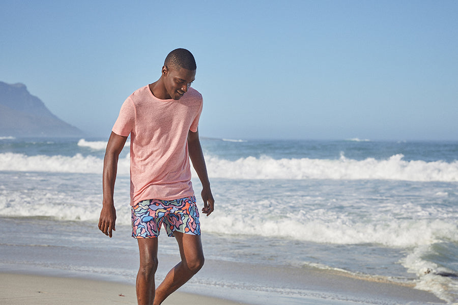 Man walking on beach in a peach t-shirt and swim shorts.