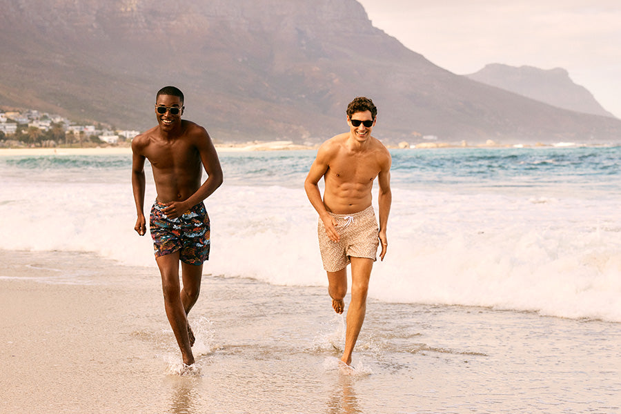Two men running on the beach in swim shorts.