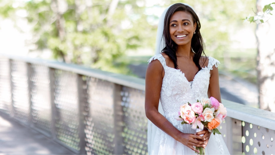 Image of bride wearing a sleeveless white lace wedding dress holding a bouquet of flowers and standing on a bridge.