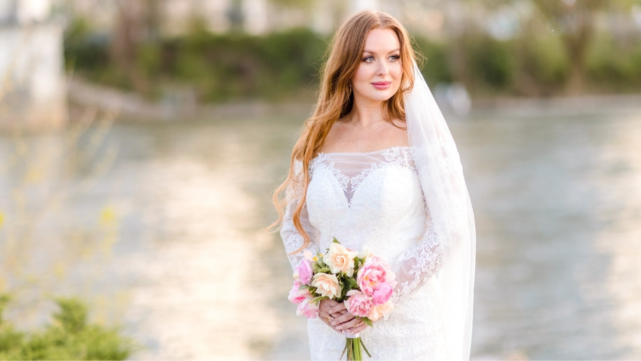Image of bride standing in front of a body of water outside wearing an off-the-shoulder white wedding dress holding a bouquet of flowers.