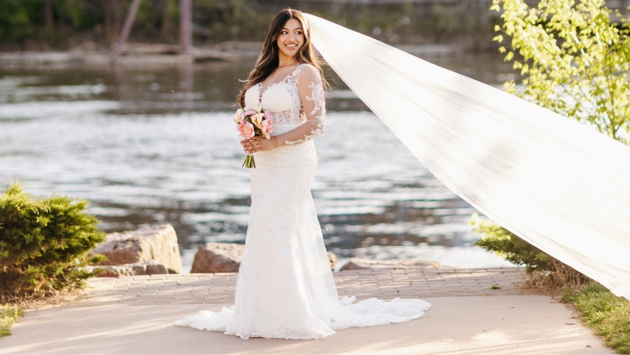 Image of a bride wearing a long white wedding dress standing at the end of a walkway in front of a body of water outside with a long veil blowing in the breeze.