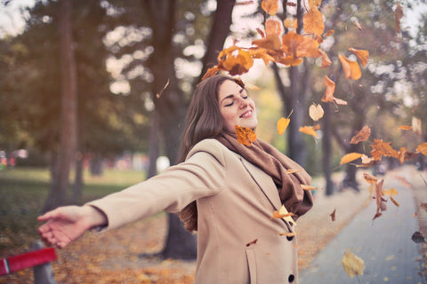 Une jeune femme brune portant un manteau beige a ses deux bras ouverts. Elle sourit et les feuilles de couleur rouge tombent sur son visage. Il y a des arbres en arrière plan.