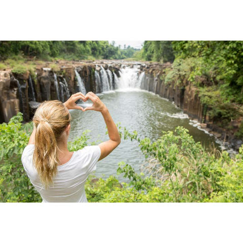 femme blonde en t-shirt blanc regarde la cascade dans la forêt et forme un cœur avec ses mains