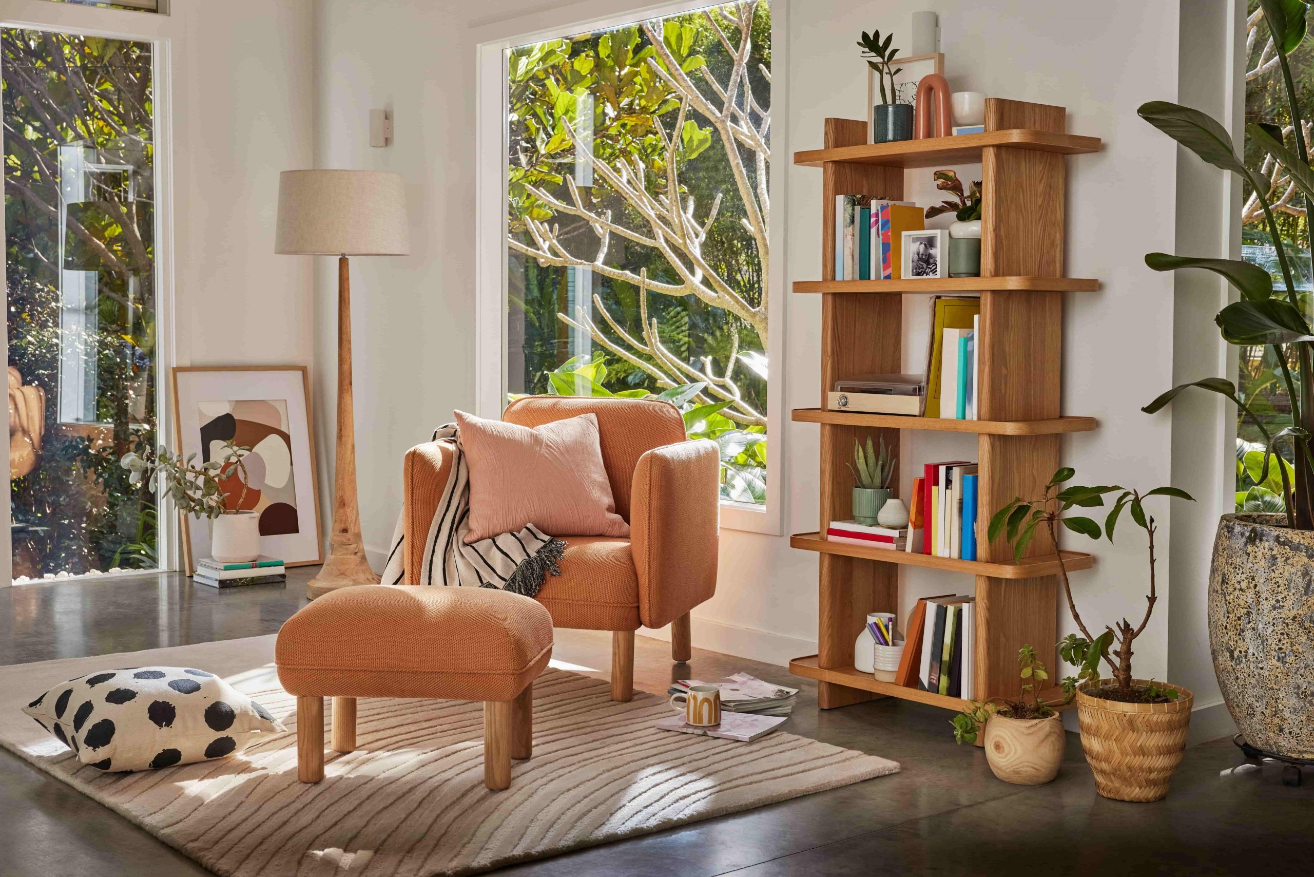 An orange single seater armchair and ottoman with a pink cushion, black and white throw rug and timber bookshelf filled with books 
