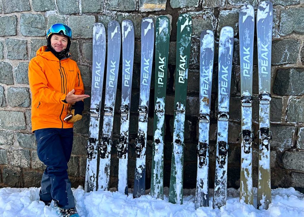 A range of Peak Skis lined up leaning on a wall in the snow
