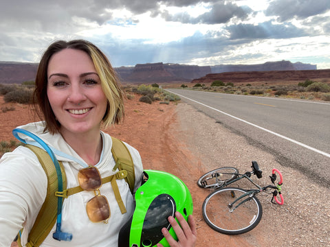 A woman stands besides a road with a bike laying down in the background.