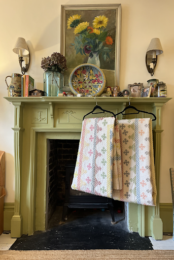 A soft green fireplace displaying an array of decorative plates, personal photographs and dried flowers. A pair of kantha quilts from Sourced by Holly hang in front.