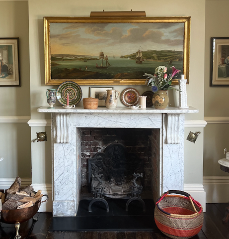 A formal drawing room marble fireplace in a Victorian home with a large antique oil painting of stormy seas. The mantlepiece is adorned with decorative plates, ceramic candleholders and earthenware jugs.