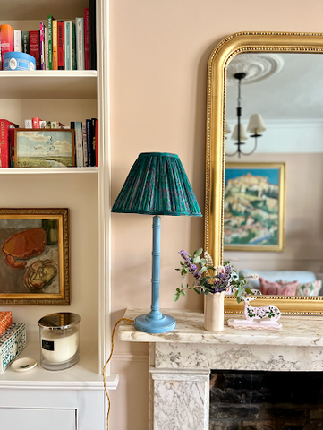A fabric lampshade placed atop a glossy bamboo lamp sits on a marble fireplace against soft pink walls. A vintage gold mirror reflects a large scenic oil painting.