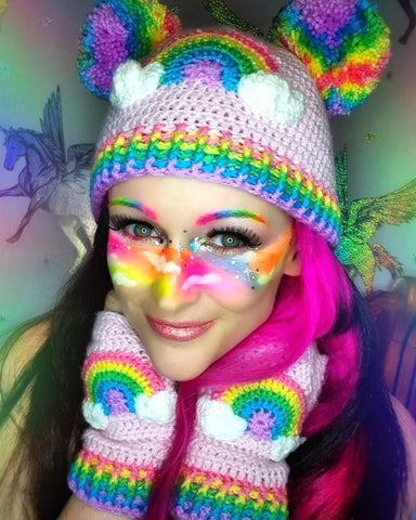 Aimee is wearing a baby pink crochet hat and gloves set, both hat and gloves feature a pastel rainbow and cloud design. Aimee also has rainbow eyebrows and makeup.