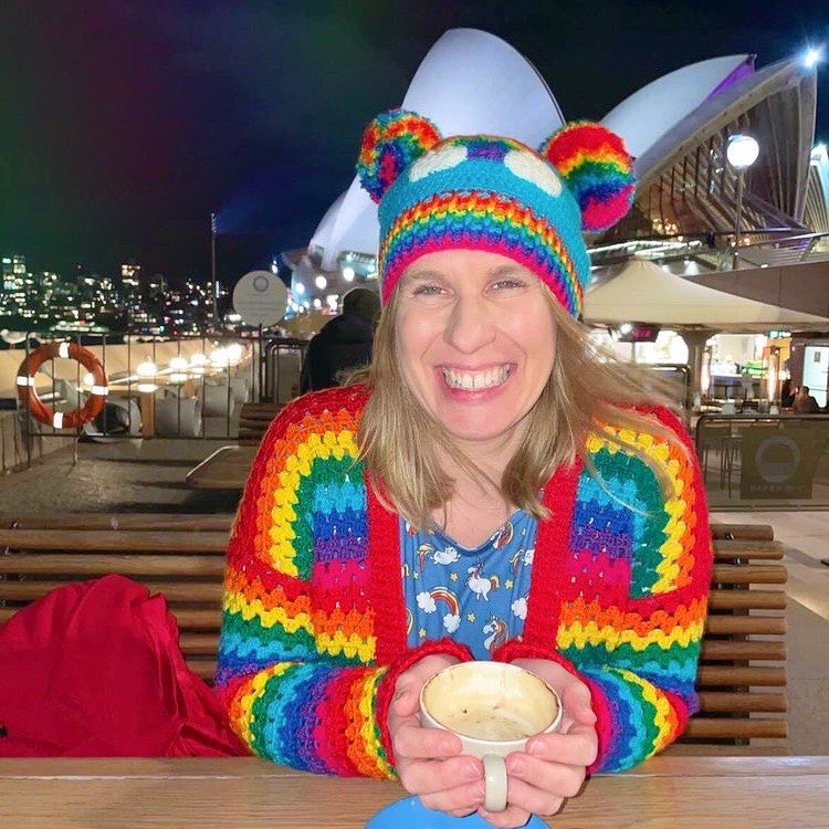 Helen, a white woman with shoulder length blonde hair is sitting on a bench at a table outside of a cafe. In the background there is a dark night sky and the Sydney Opera can be seen behind her. Helen is holding a cup of what seems to be coffee with both hands and has a wide smile on her face. Helen is wearing a blue dress with rainbow cloud print, a crocheted rainbow striped cardigan and a turquoise crocheted hat with rainbow pom pom 'ears', a rainbow cloud motif on the front and a rainbow striped ribbed brim.