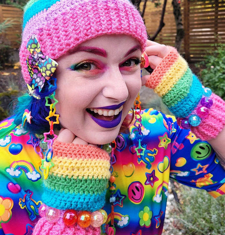 Vala who is dressed in very colourful clothing with rainbow eyeshadow, pink eyebrows and purple lipstick, is wearing the VelvetVolcano Pastel Rainbow Striped Pom Pom Beanie and Pastel Rainbow Striped Fingerless Gloves