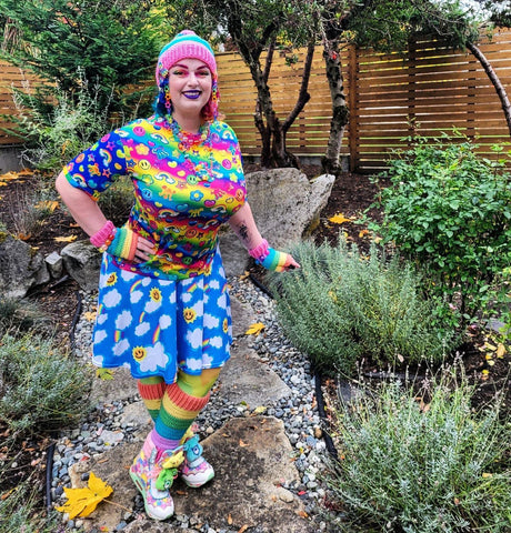 Vala who is dressed in very colourful clothing with rainbow eyeshadow, pink eyebrows and purple lipstick, is wearing the VelvetVolcano Pastel Rainbow Striped Pom Pom Beanie, Pastel Rainbow Striped Fingerless Gloves and Pastel Rainbow Striped Leg Warmers.