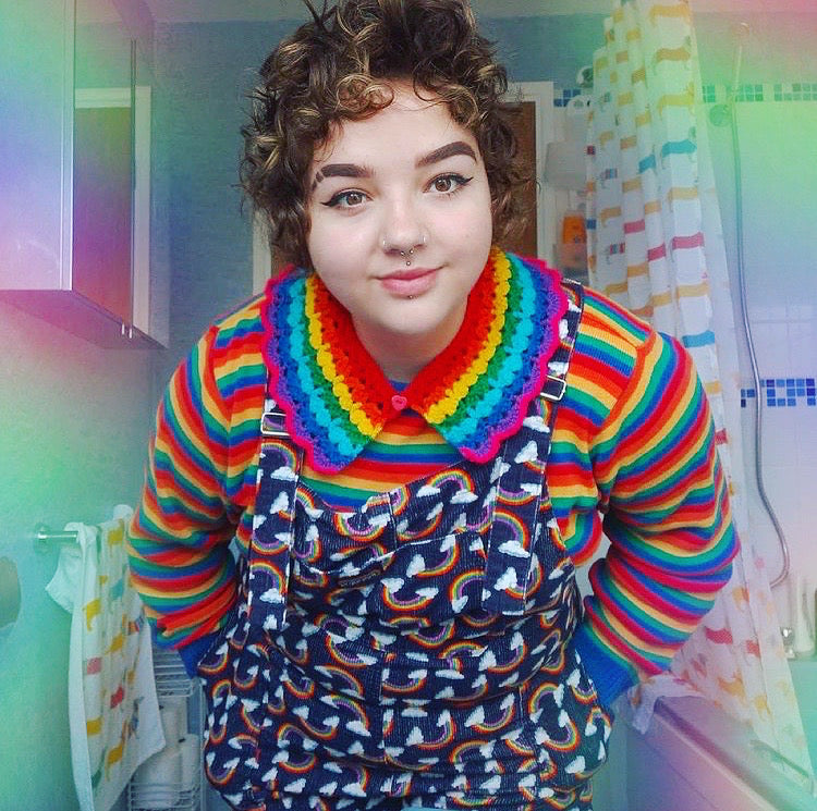 Samantha wearing a crocheted rainbow striped collar, rainbow striped long sleeve t-shirt and a pair of navy dungarees with a rainbow cloud print.