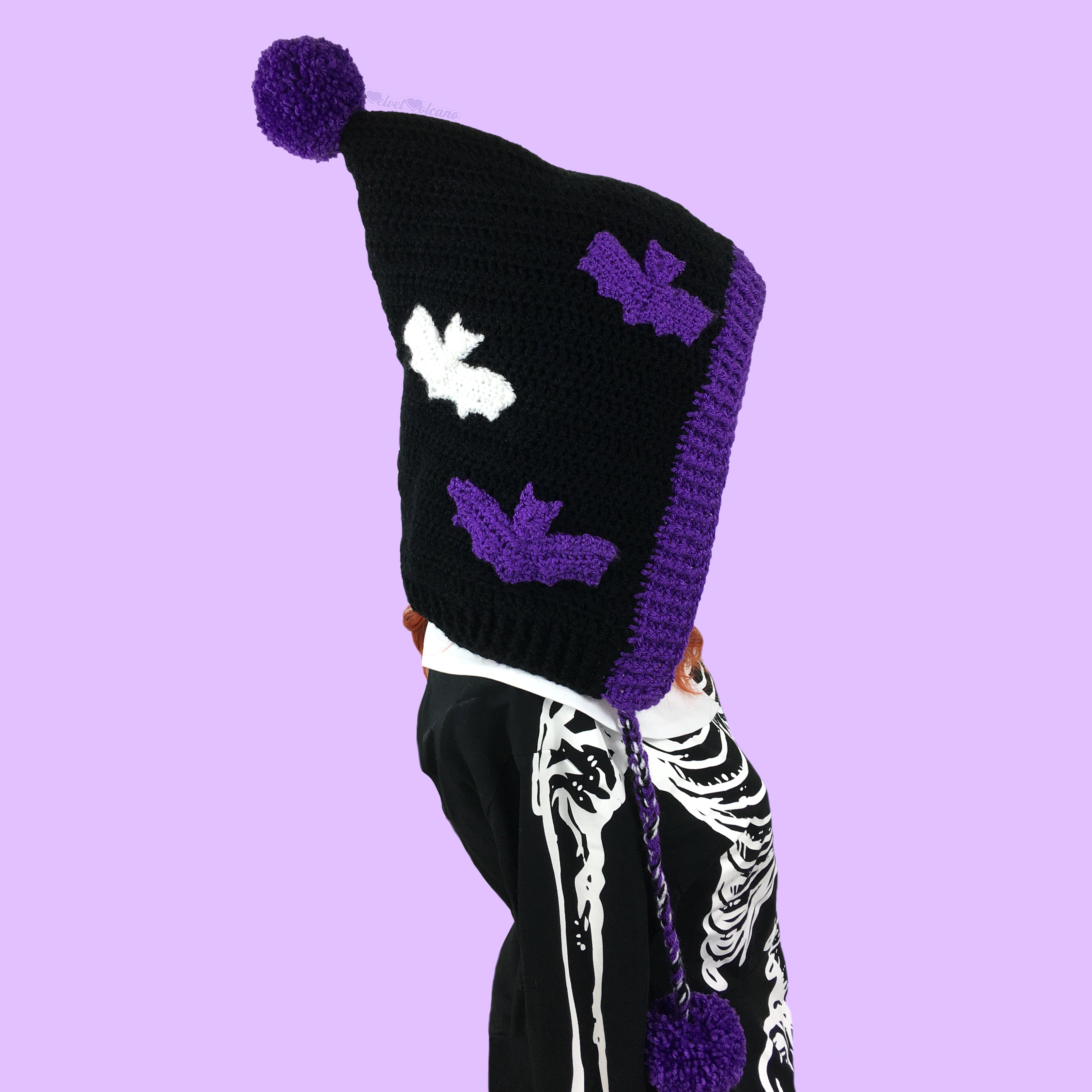 A black crocheted pixie hood with purple and white bat applique pattern, purple trim, ties and a purple pom pom at the point of the hood.