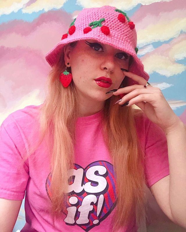 Brooke is wearing a bubblegum pink crocheted bucket hat with red and green cherries on it, Brooke is also wearing a bright pink T-shirt that says “as if!” in reference to the 90’s cult film Clueless