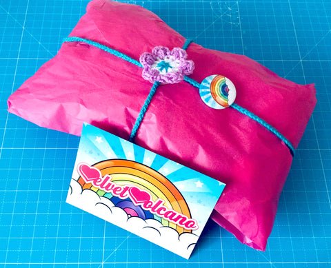 A pink tissue paper wrapped parcel with a lilac, turquoise and white crocheted flower tie wrapped around it. Attached to the tie is a rainbow badge and in front of the parcel is a VelvetVolcano rainbow flyer. The background is a turquoise fabric cutting board.