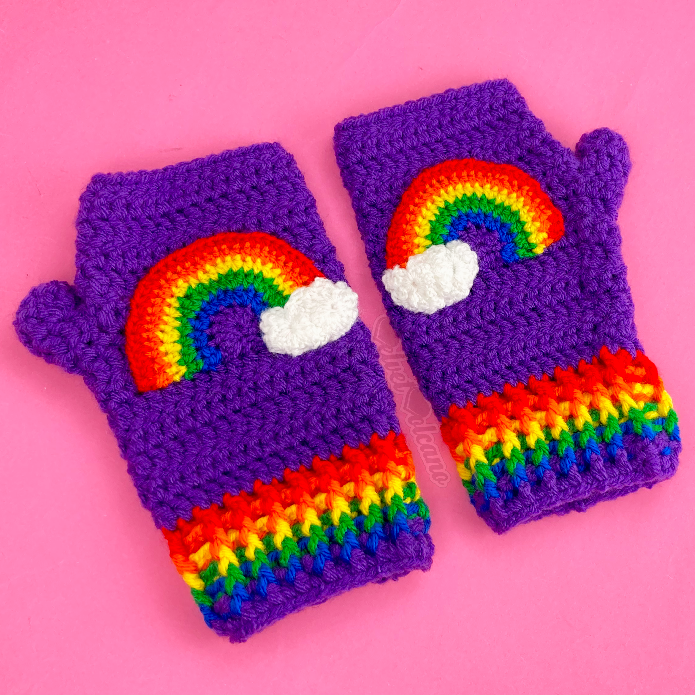 A purple pair of crocheted fingerless gloves with a rainbow striped cuff and a rainbow applique on the front of each glove that has a white cloud on one end of the rainbow.