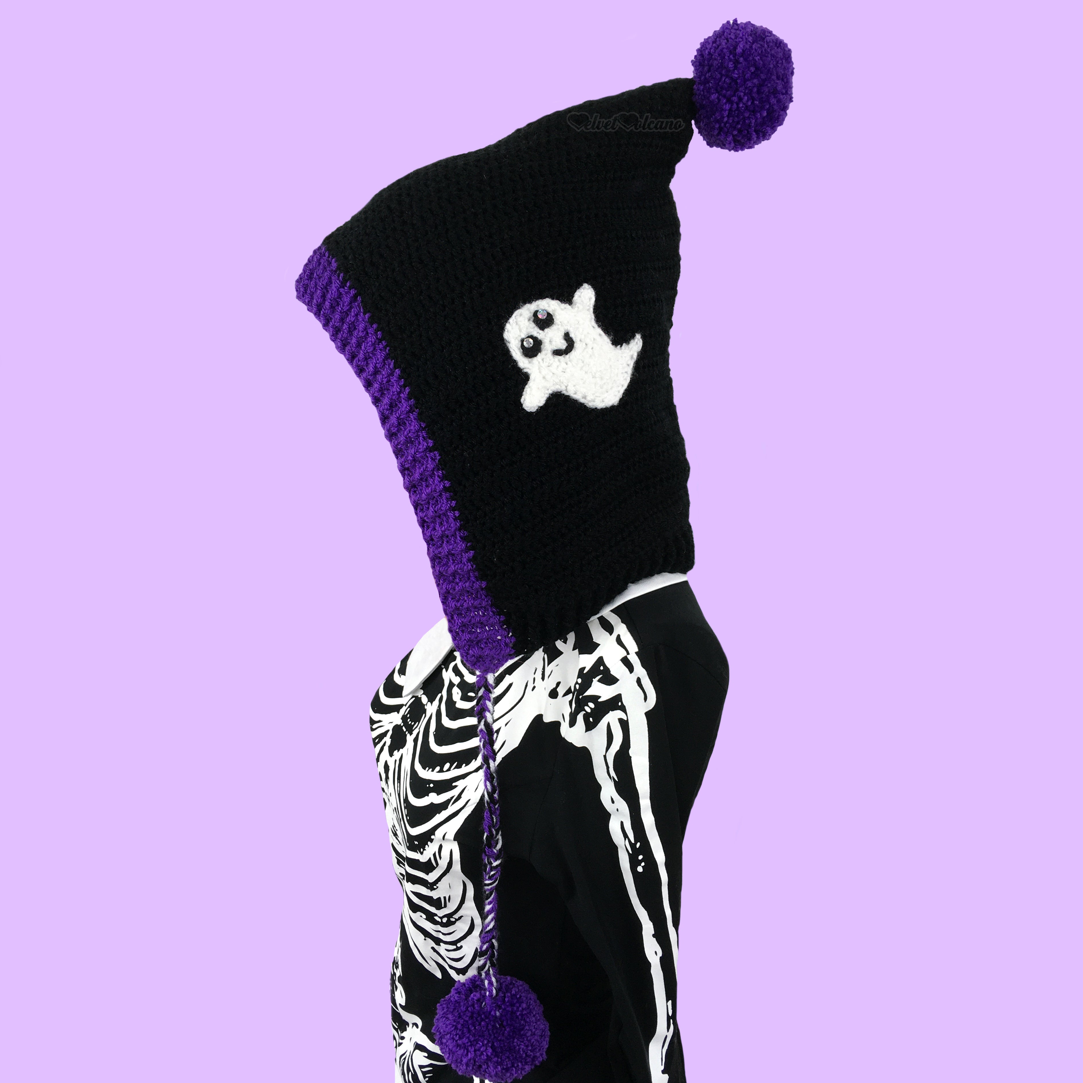 A black crocheted pixie hood with white ghost applique, purple trim, ties and a purple pom pom at the point of the hood.