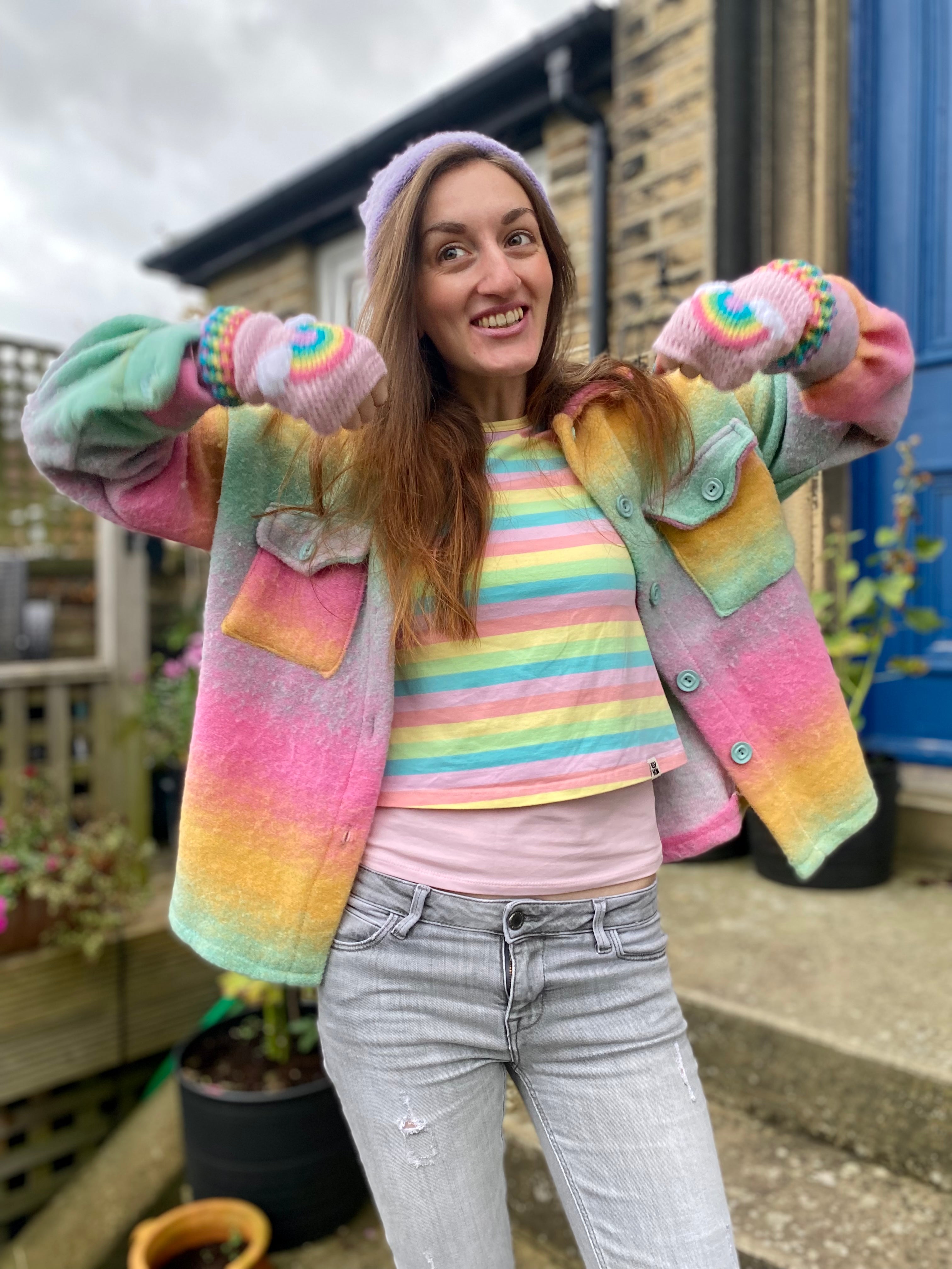 Rachel, a woman with long brown hair is wearing a pastel rainbow striped T-shirt, a pastel rainbow ombré shacket, a lilac beanie and VelvetVolcano baby pink crocheted fingerless gloves with pastel rainbow and cloud motif and pastel rainbow striped gloves.