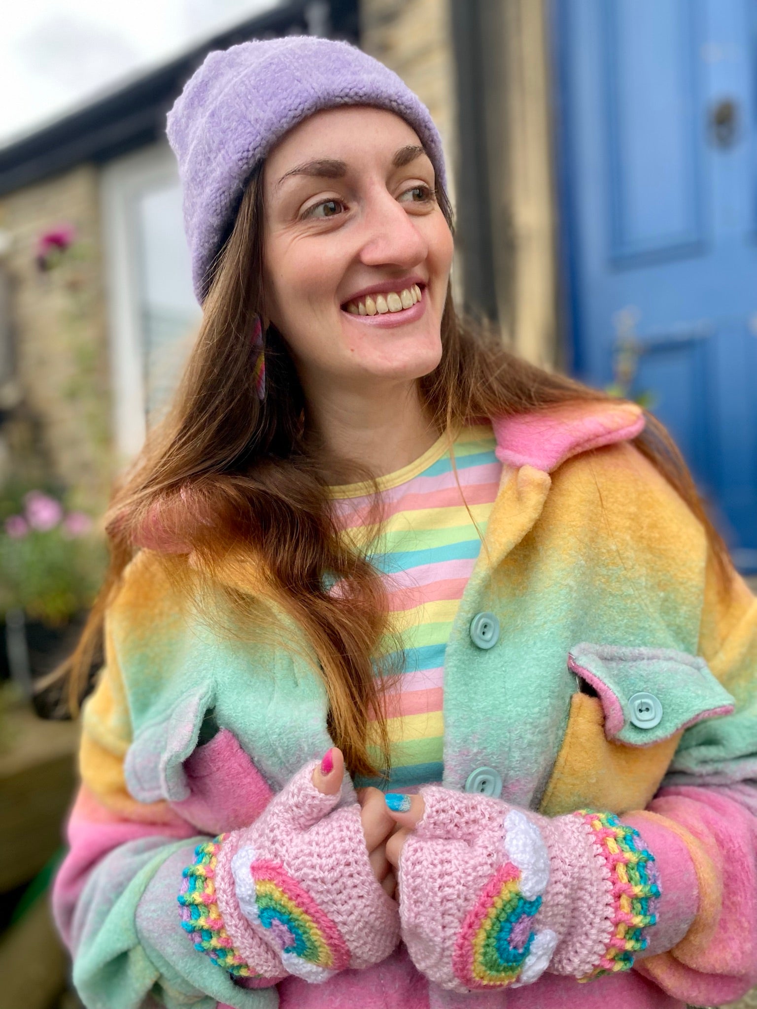 Rachel, a woman with long brown hair is wearing a pastel rainbow striped T-shirt, a pastel rainbow ombré shacket, a lilac beanie and VelvetVolcano baby pink crocheted fingerless gloves with pastel rainbow and cloud motif and pastel rainbow striped gloves.