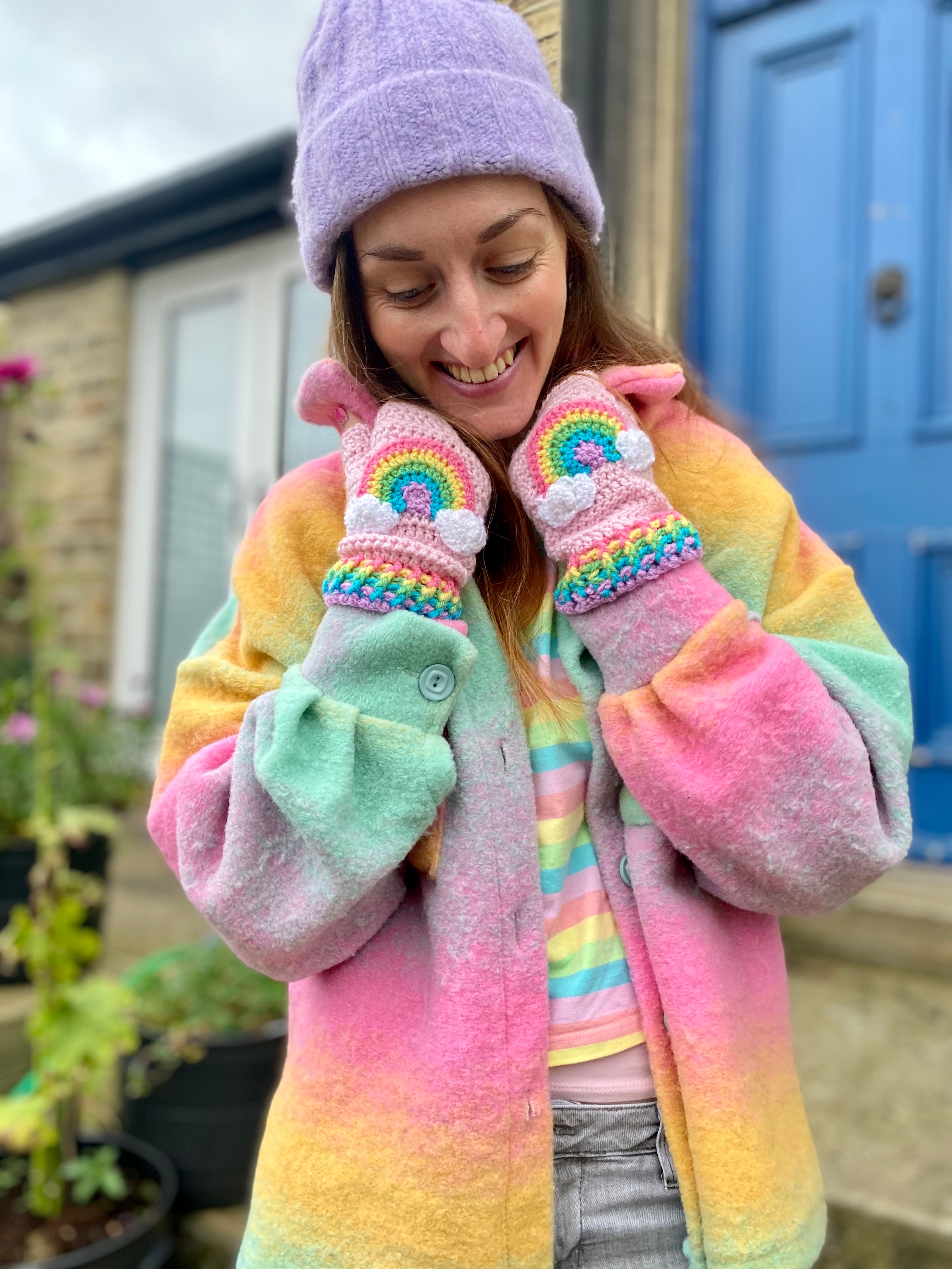 Rachel, a woman with long brown hair is wearing a pastel rainbow striped T-shirt, a pastel rainbow ombré shacket, a lilac beanie and VelvetVolcano baby pink crocheted fingerless gloves with pastel rainbow and cloud motif and pastel rainbow striped gloves.