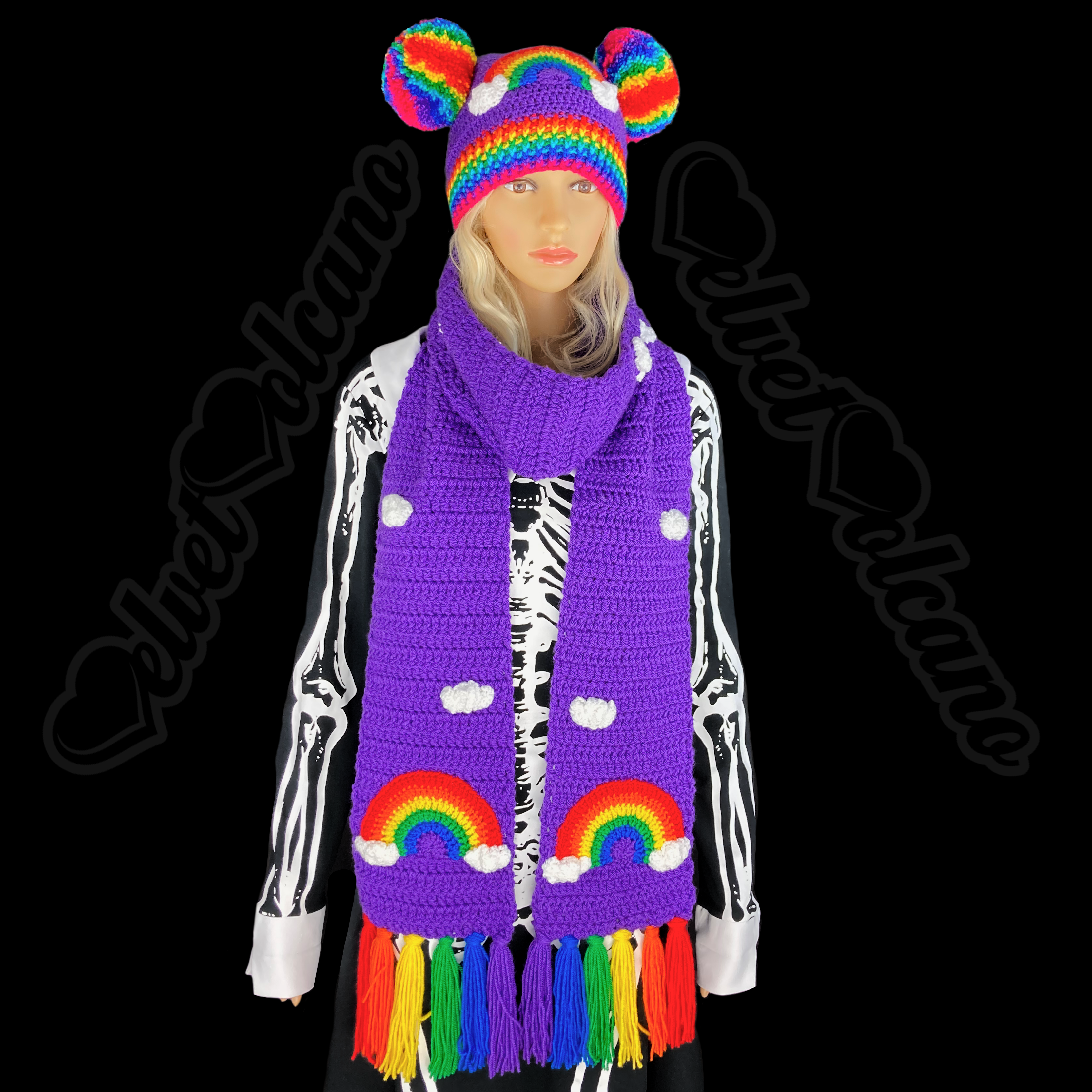 A purple crocheted beanie and scarf set. The beanie features 2 rainbow pom poms at the top of the hat, positioned like animal ears, a rainbow applique with white clouds on either end and a rainbow striped brim. The scarf features a repeating white cloud applique pattern and a rainbow cloud applique on each end of the scarf, the scarf also features rainbow tassels.