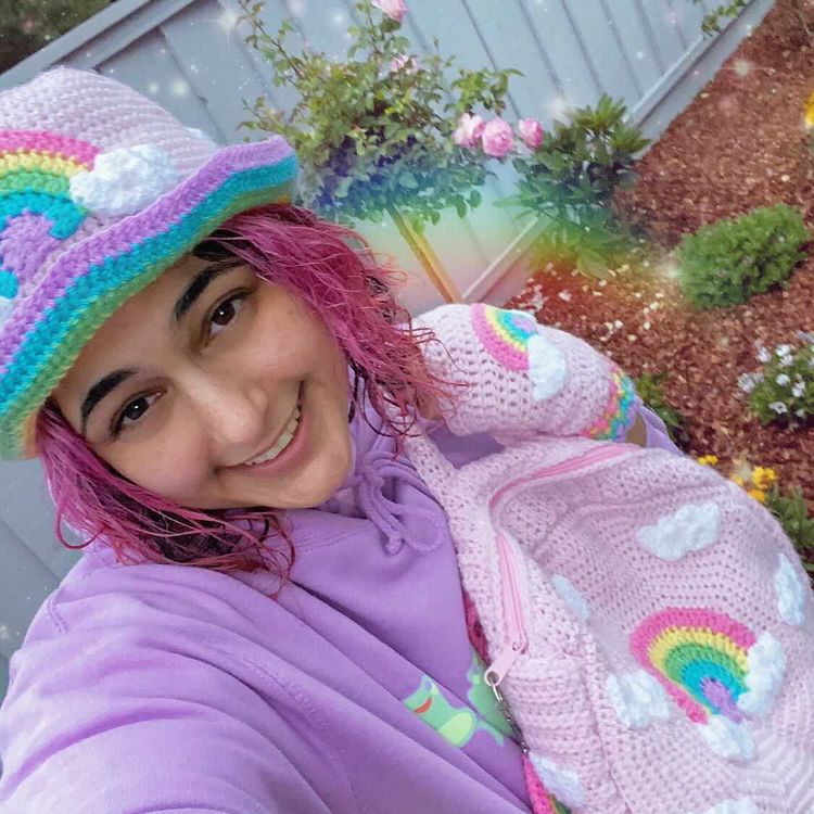 Sabrina who has short pink hair is wearing a lilac hoodie and 3 VelvetVolcano crocheted items including a Pastel Rainbow Cloud Bucket Hat, a pair of Pastel Rainbow Cloud Fingerless Gloves and a Pastel Rainbow Cloud Backpack. 