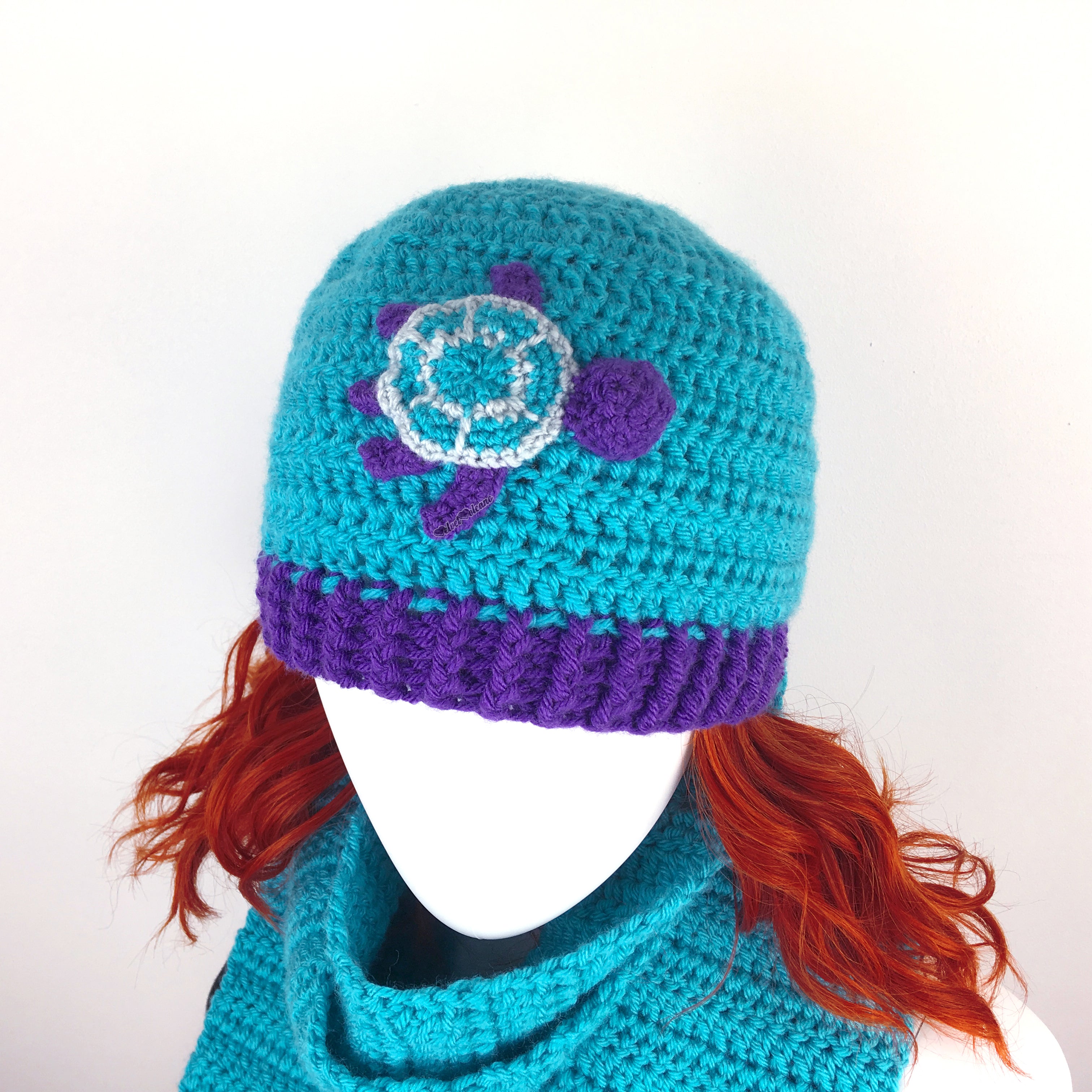 A turquoise crocheted beanie with purple brim and a turquoise, purple and light grey turtle applique on the front.