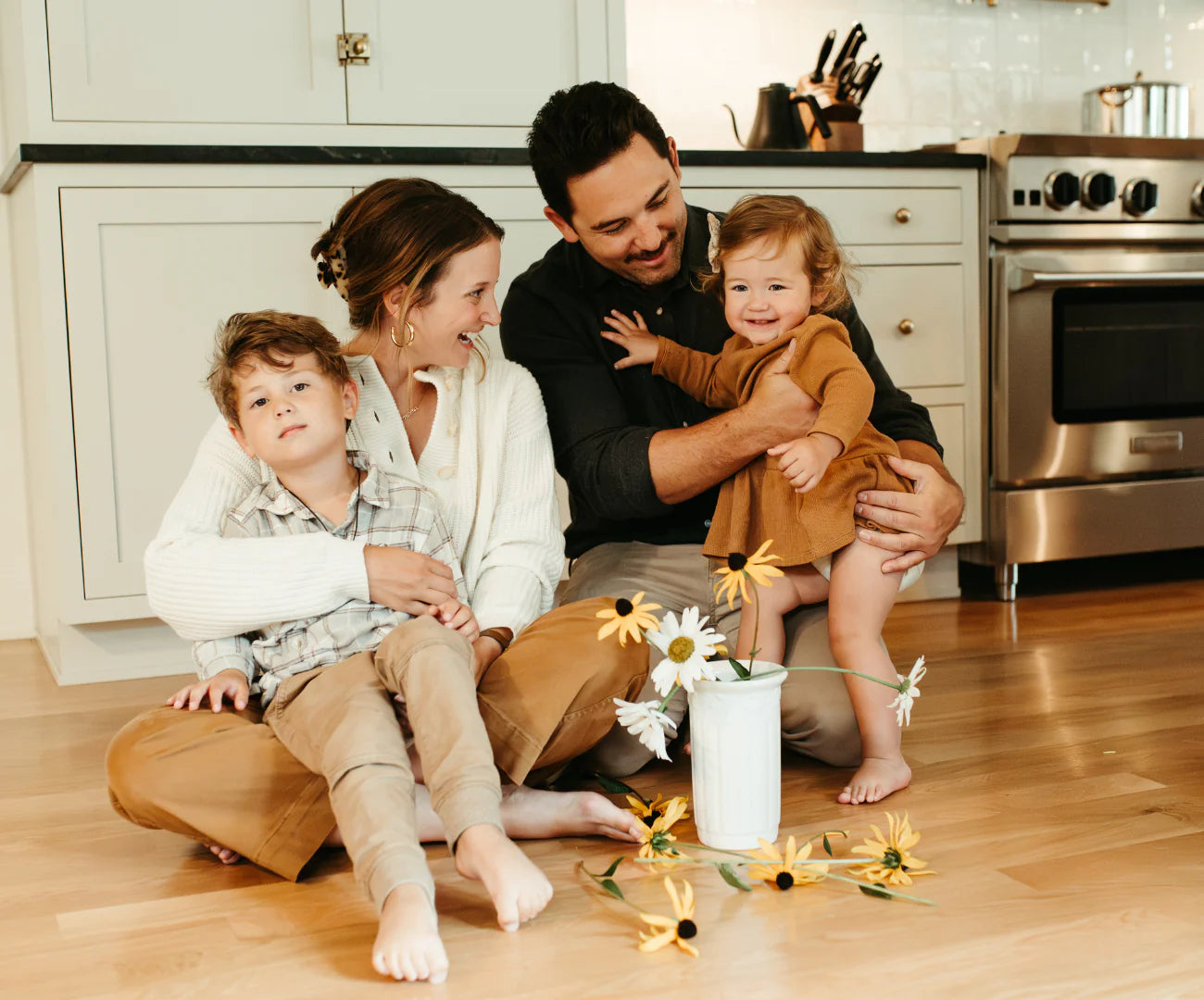 A happy family with two children sitting on the kitchen floor, surrounded by scattered flowers.