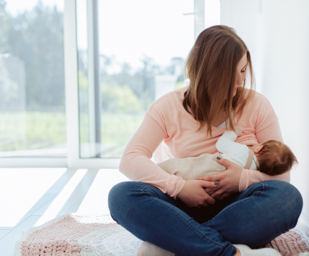 a mom sitting cross-legged on the floor nursing her newborn baby postpartum