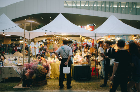 a malaysian-style bazaar. white tents with warm/yellow fairy lights brighten the tents as a mass of people shop and walk about.