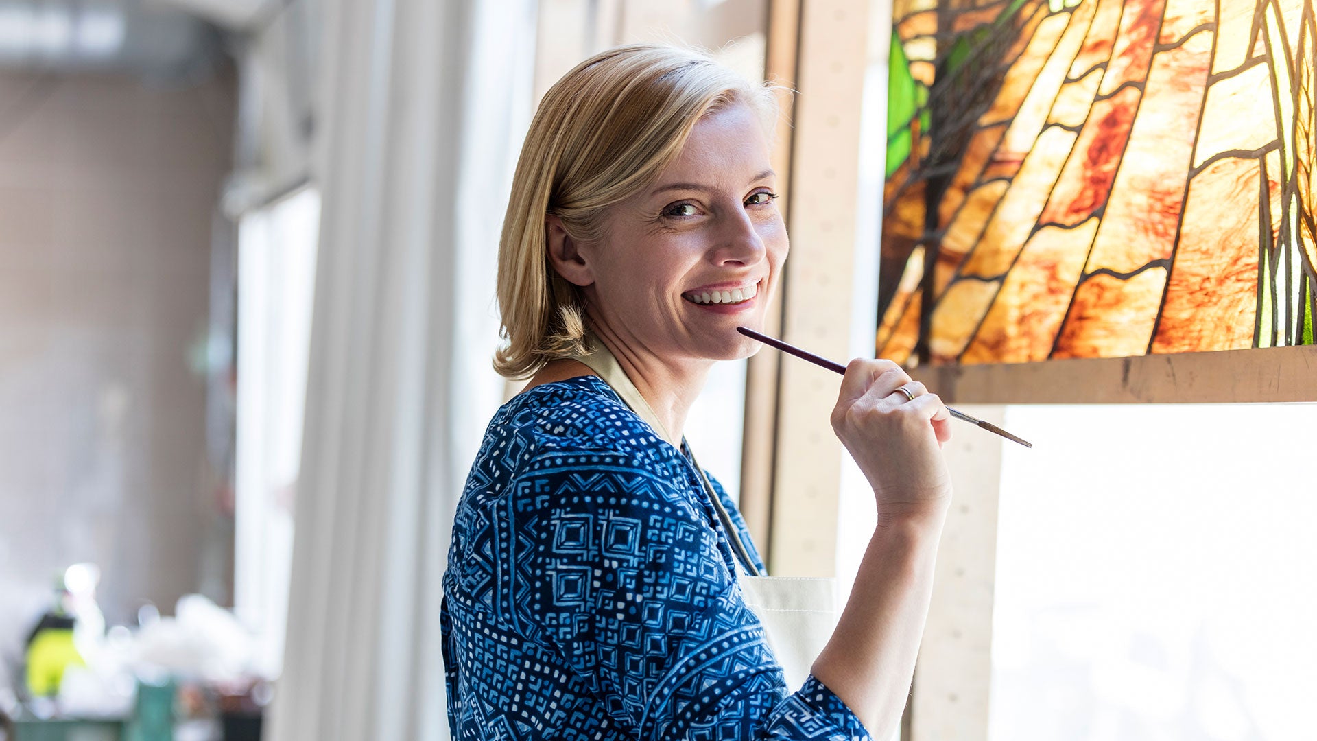 WOman looking at camera with finished stained glass project displayed in background.