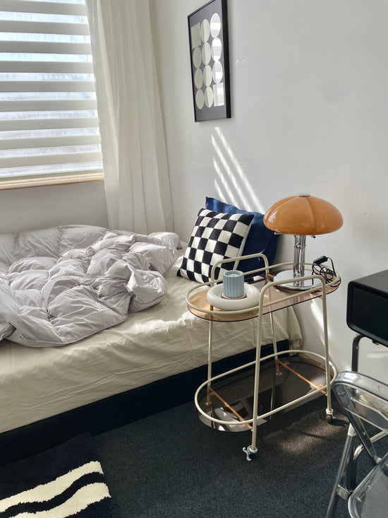 A  stylish bedside setup featuring a small bar cart with a retro orange lamp, a modern candle in a ceramic holder, and decorative accents. 