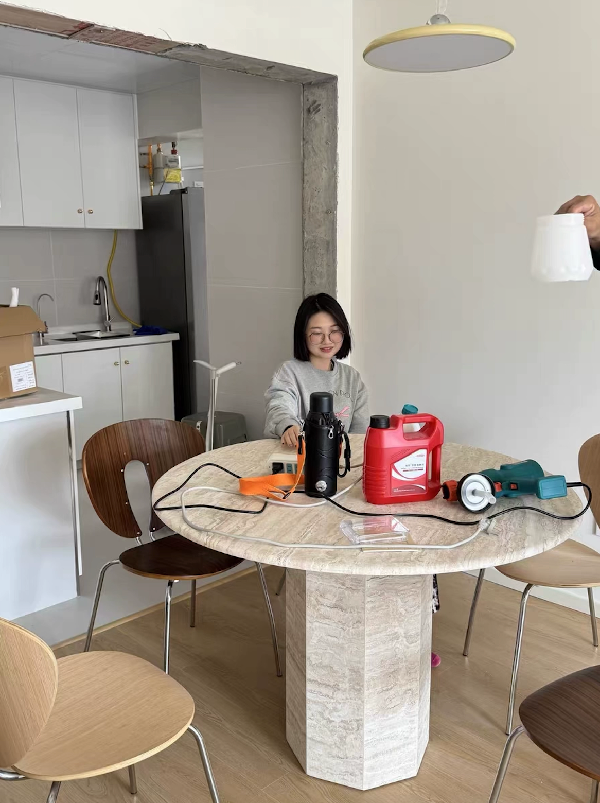 A modern dining area featuring a round travertine table with natural stone texture, surrounded by wooden chairs. A woman is seated at the table, which is temporarily being used for tools and supplies, creating a casual and informal scene. In the background is a minimalist kitchen with white cabinets.