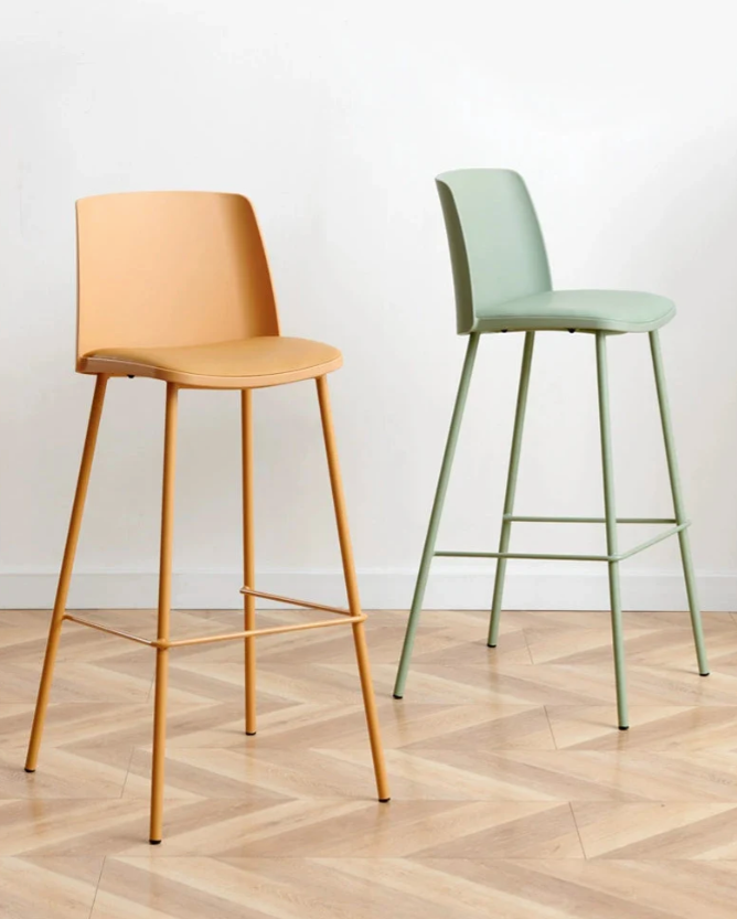 Two modern bar stools against a white background on a herringbone patterned wood floor. The stool on the left features a warm peach seat and matching metal frame, while the one on the right has a pastel green seat and a green metal frame. Both stools have a sleek, minimalist design with slender legs and a subtle footrest.