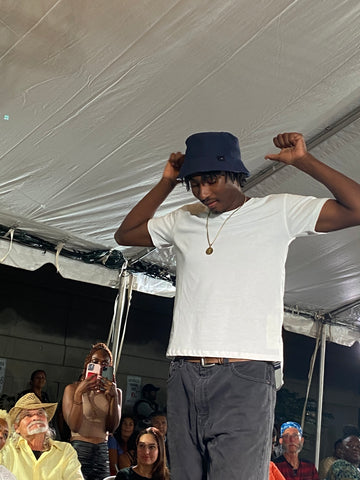 man in white tee wearing navy bucket hat pointing with his thumbs toward his back