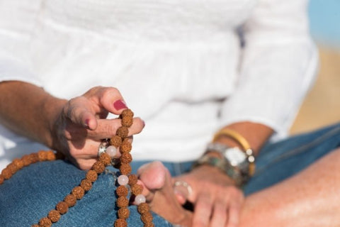 Woman in white top and jeans sat crossed legged on beach holding a mala in her hand between her middle finger and thumb