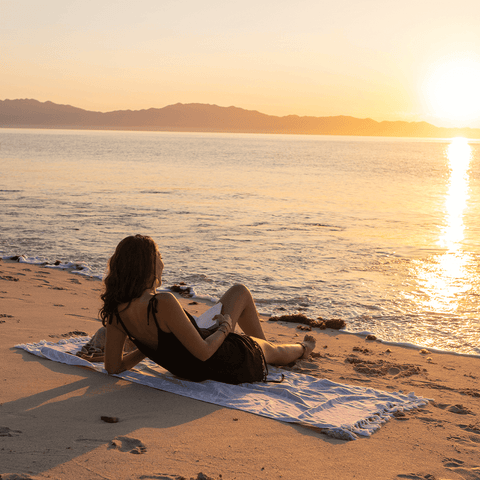 A woman laying on a Turkish towel at the beach watching the sunset. 