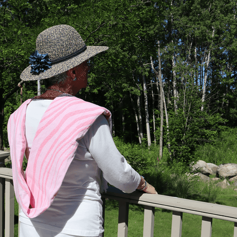 Elderly woman draped in a folded pink patterned Turkish towel, looking outside on a sunny day with a hat on.