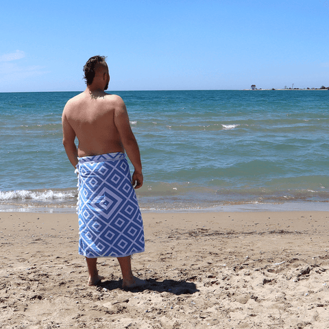 Man standing on a beach wrapped in a blue diamond Pomp & Sass Turkish towel, looking off into the distance.