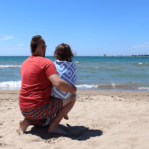 A father and a child looking out on the beach, the child is wrapped in a blue diamond patterned Turkish towel.