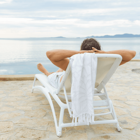 Man laying down on a bench on the beach looking out into the water, a white Pomp & Sass Turkish towel is hanging on the chairs end.