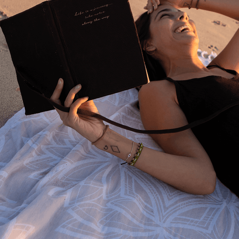 Woman at a beach laying on her back on top of a cream coloured Turkish towel, reading a book.