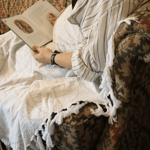 Woman sitting on couch under a white Turkish towel reading a book on a patterned couch.