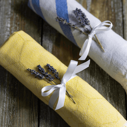 One yellow and one white/blue Turkish towel on a wooden surface wrapped with a white satin bow and sprigs of lavender for gifitng