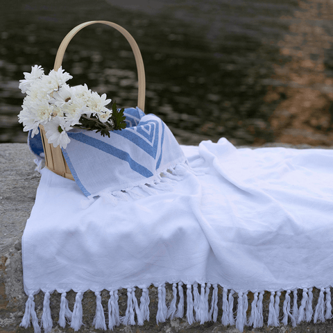 A white Turkish towel. used as a picnic blanket by the lake. With a brown picnic basket that holds white flowers and a blue diamond Turkish towel.