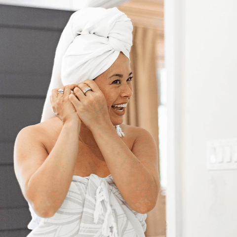 Smiling woman in a grey and white Turkish towel used as a sarong in the bathroom. She is drying her hair with a white Turkish towel.